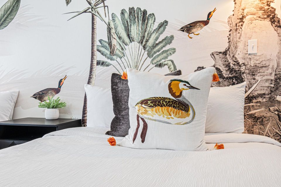 Bed with bird-decorated pillow, against a wall mural of tropical plants and birds.