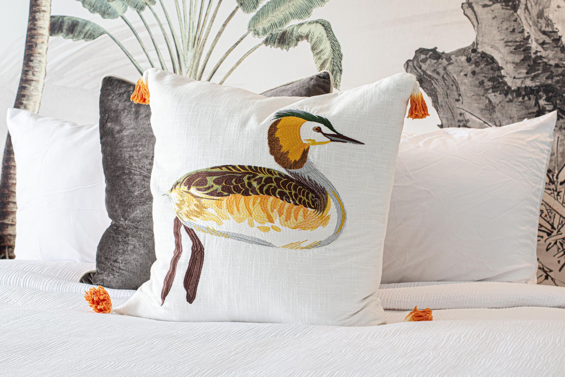 White bird-embroidered pillow with orange tassels, on a bed with a tropical-themed wallpaper background.