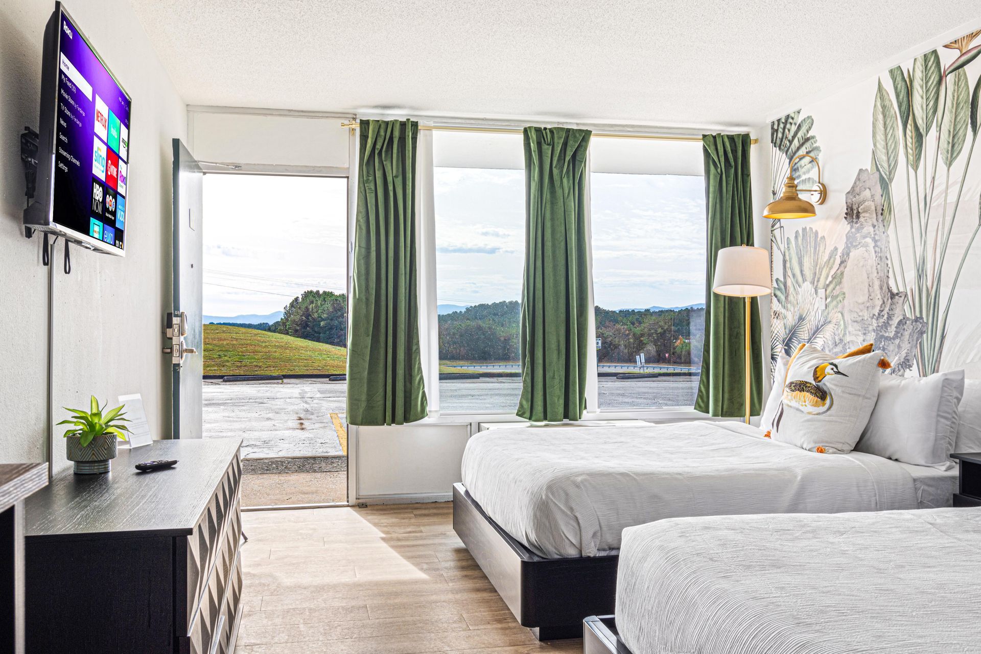 Hotel room with two beds, large window with green curtains, and a view of a green landscape.