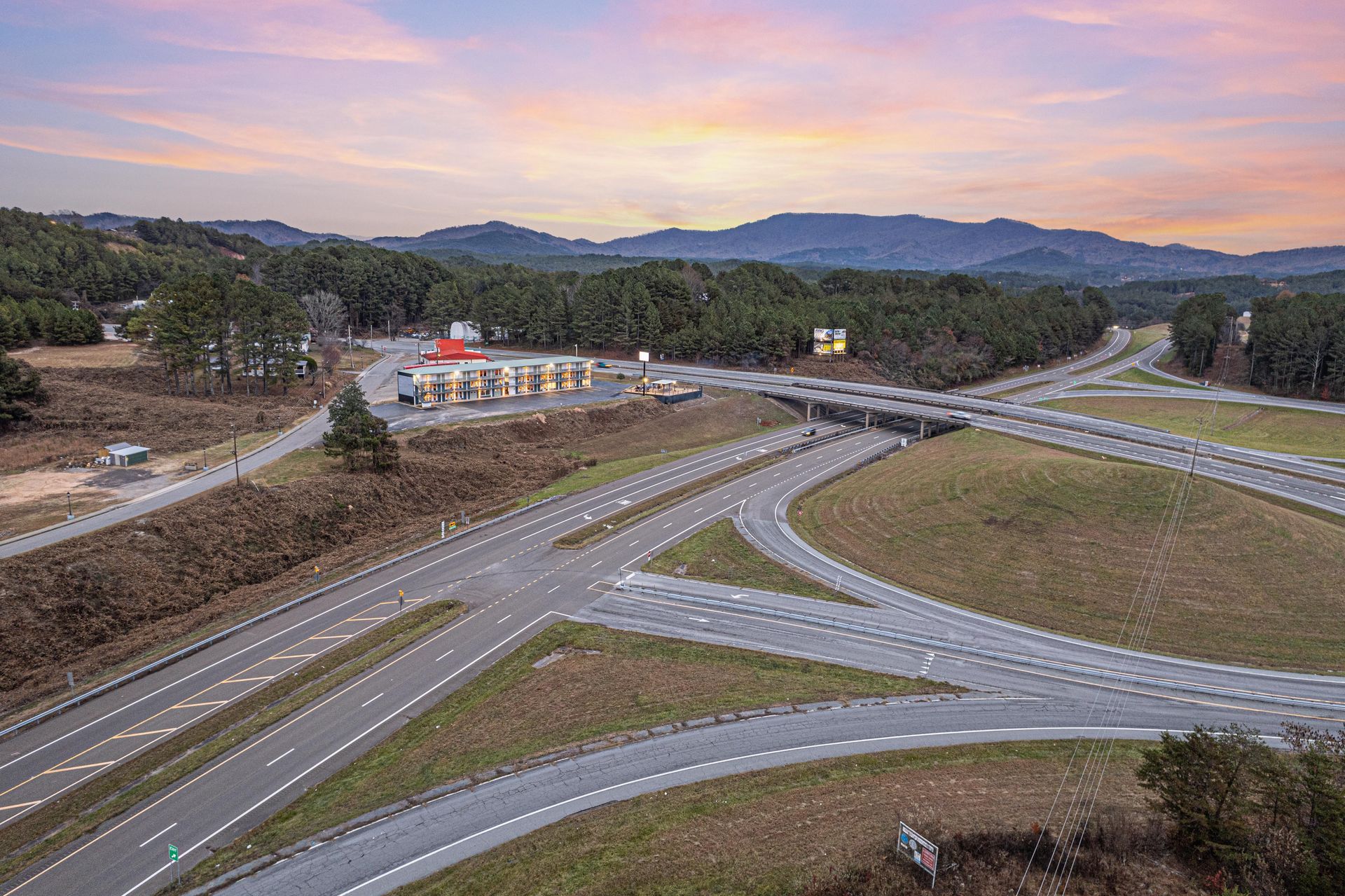 Highway interchange with roads, overpass, gas station, and forested hills at sunset.