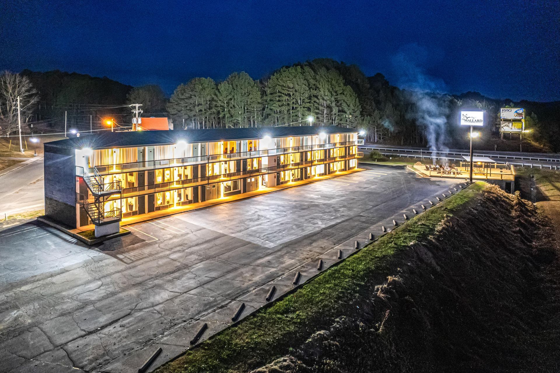A motel at night with illuminated rooms, a parking lot, and a small fire pit, located near a road and trees.
