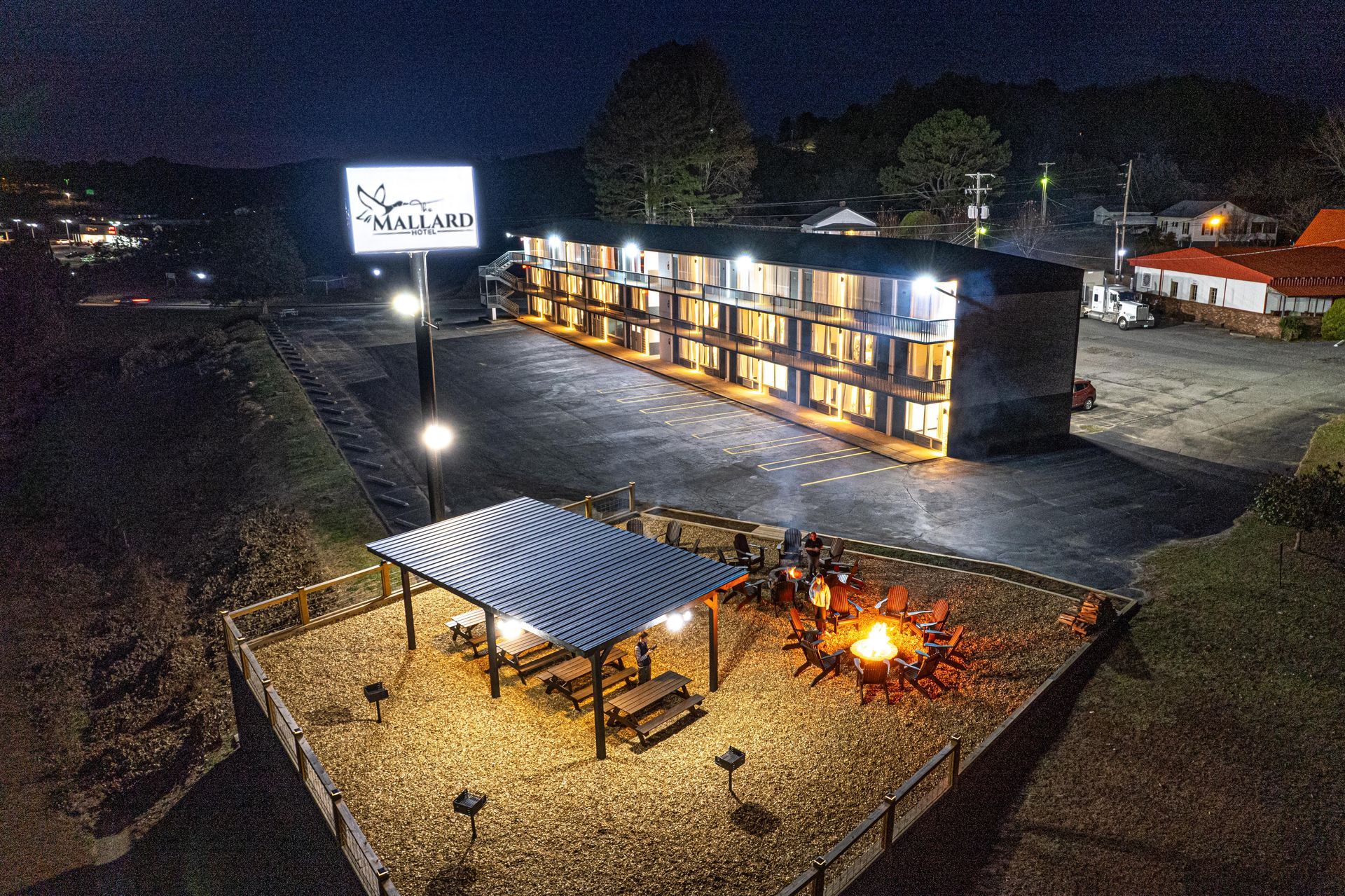 Night aerial view of a motel with illuminated sign, surrounded by parking, a fire pit, and a covered seating area.