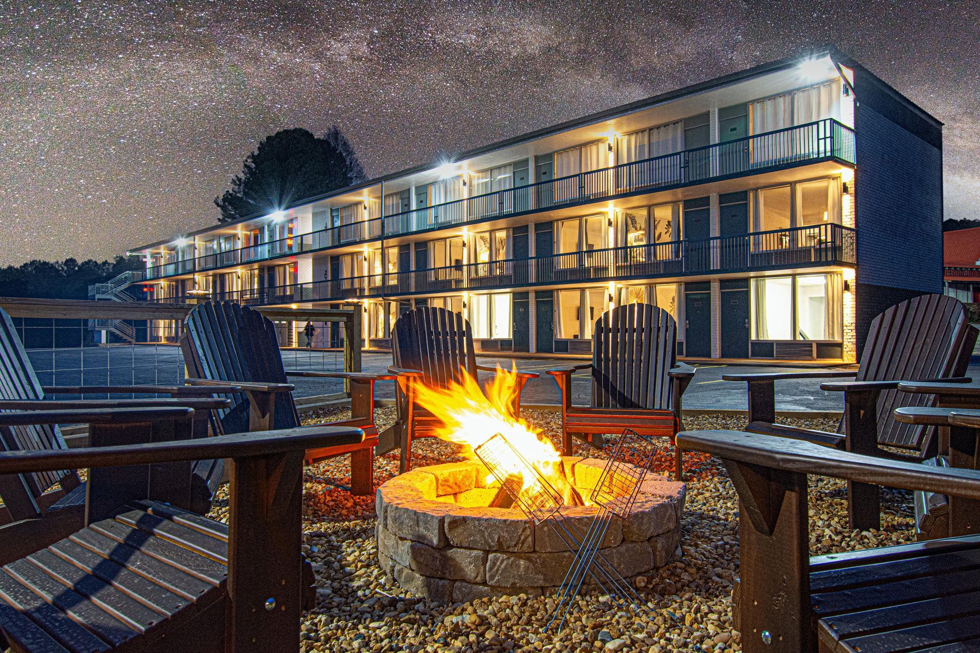 A motel at night with a lit bonfire and Adirondack chairs under a starry sky.