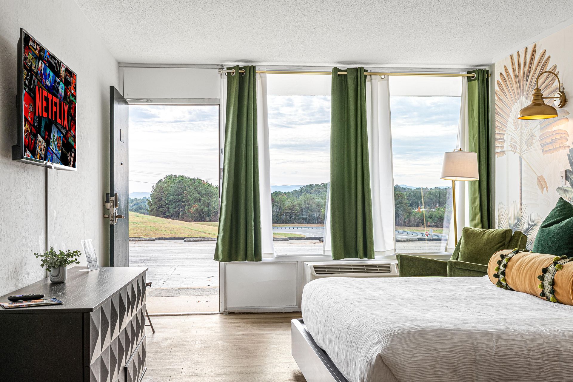 Hotel room with bed, TV, open door to outdoor view, green curtains.