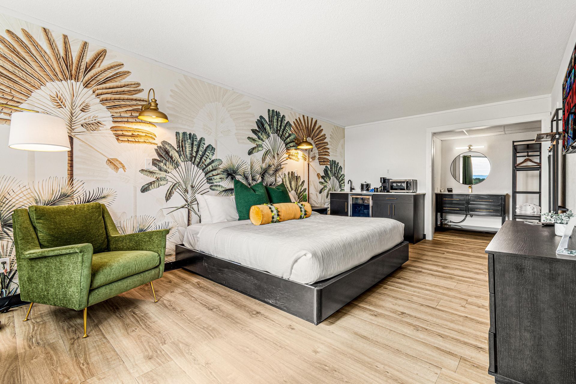 Hotel bedroom with a king-sized bed, green armchair, palm tree wallpaper, and hardwood floors.