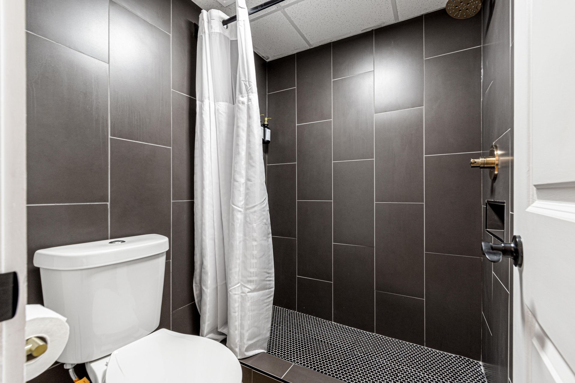 A small bathroom with a toilet and a tiled shower with a white shower curtain.