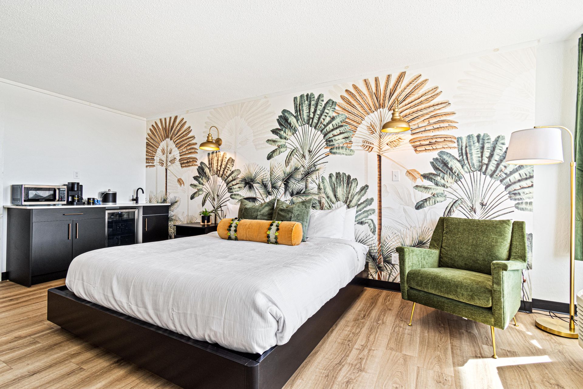 Hotel bedroom with bed, green armchair, palm-tree wallpaper, and mini fridge.