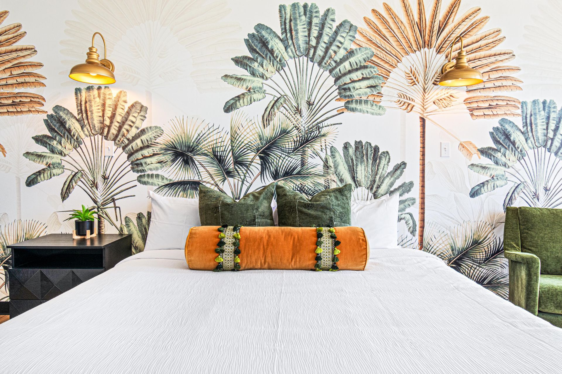 Bed with tropical mural headboard, orange bolster, green pillows, brass sconces, and green velvet chair.