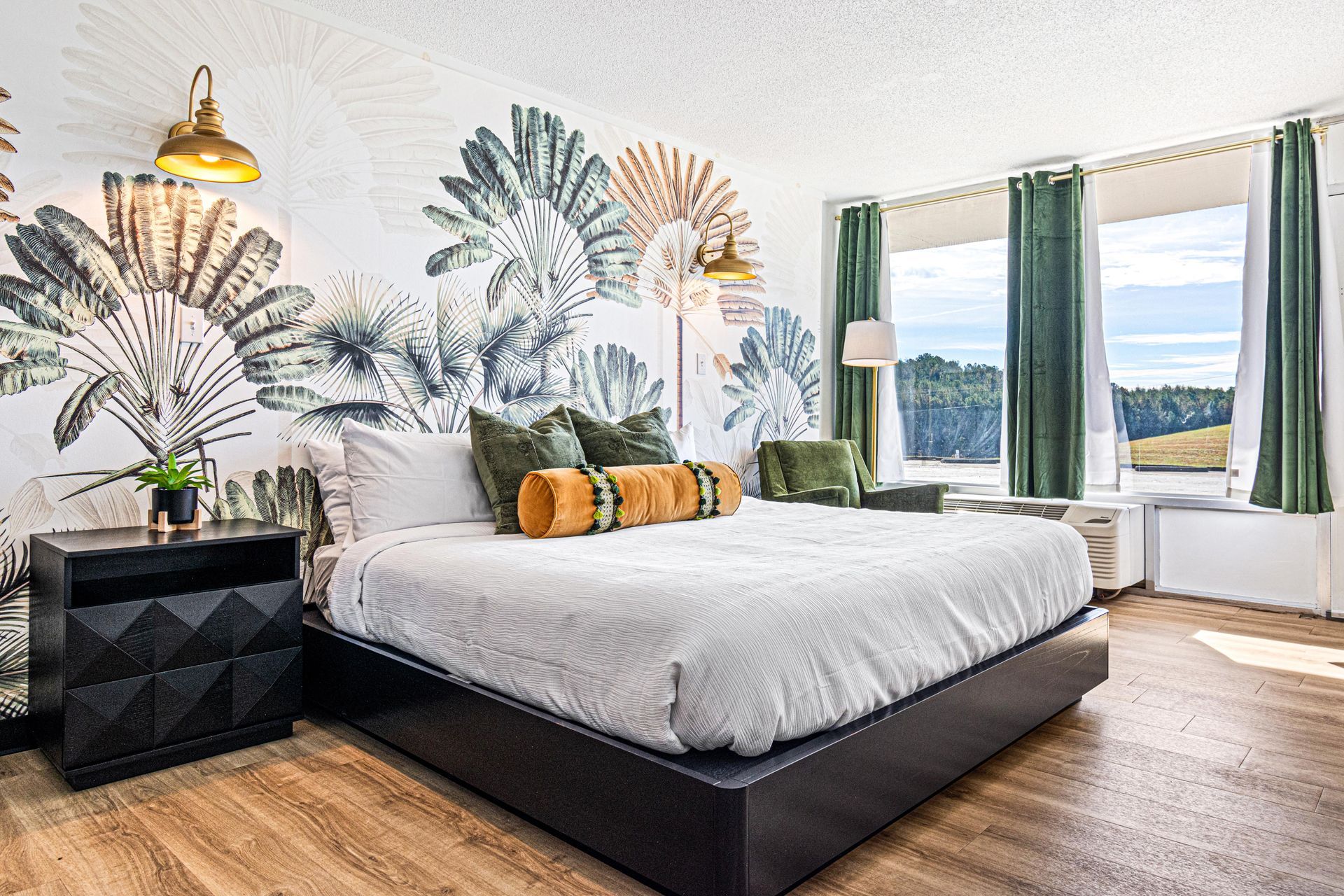 Bedroom with a palm leaf mural, bed with green and gold pillows, gold sconces, and a scenic view.