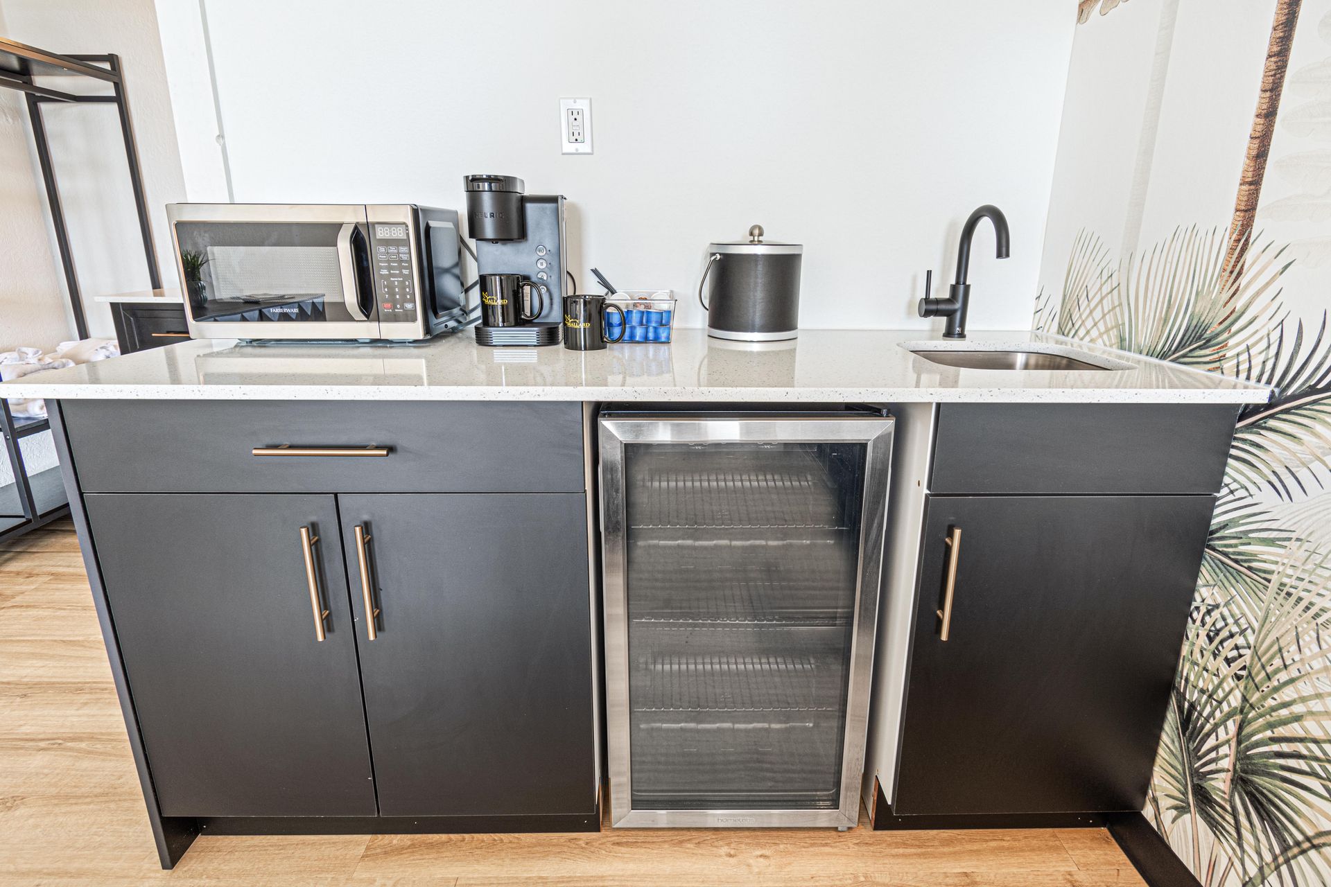 Small kitchenette with microwave, coffee maker, sink, and wine cooler; black cabinets and white countertop.