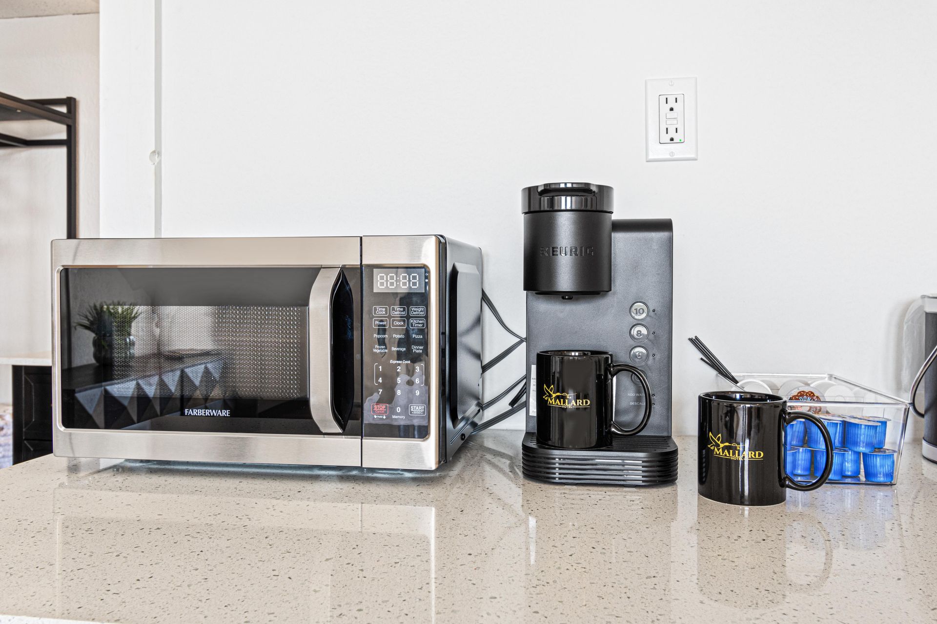 Microwave, coffee maker, mugs, and snacks on a countertop against a white wall with an electrical outlet.