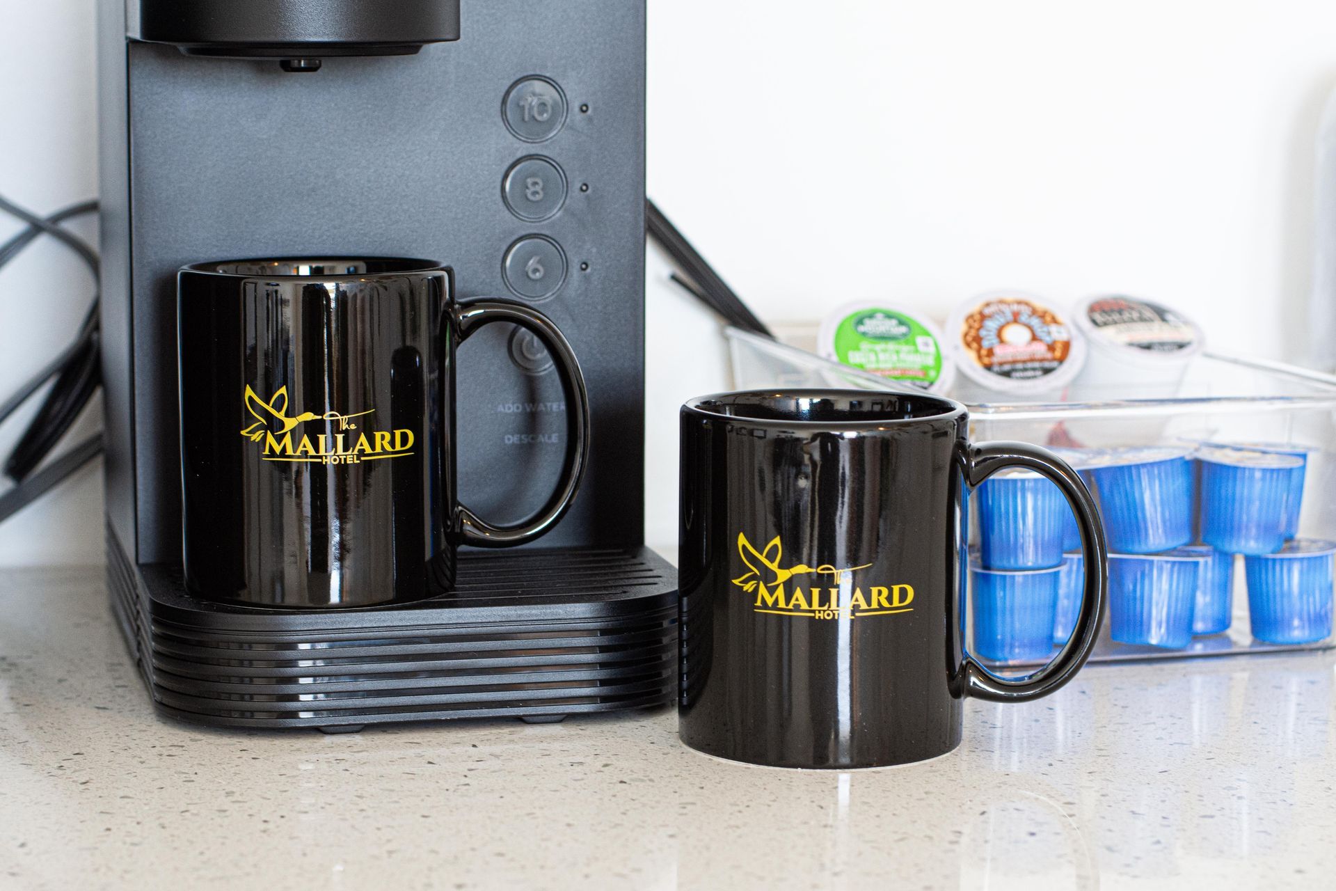 Two black mugs with a gold logo and a coffee machine on a countertop.