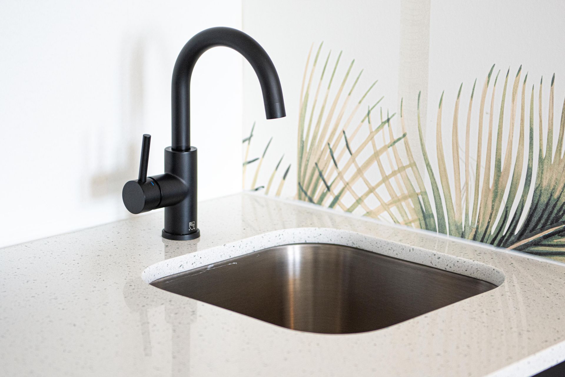 Black faucet over a stainless steel sink in a light-colored countertop, with a palm leaf patterned backsplash.