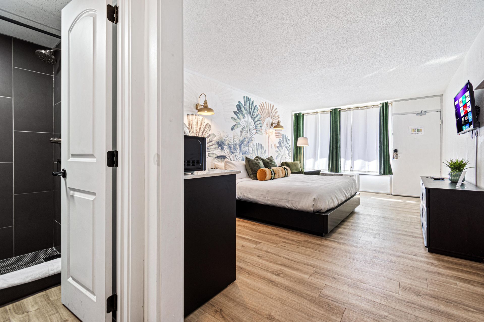 Hotel room with a bed, TV, bathroom, and floral accent wall; light wood flooring.
