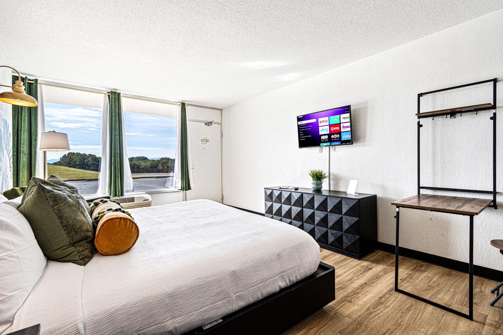 Hotel room with bed, TV, dresser, clothing rack, and large window overlooking water.
