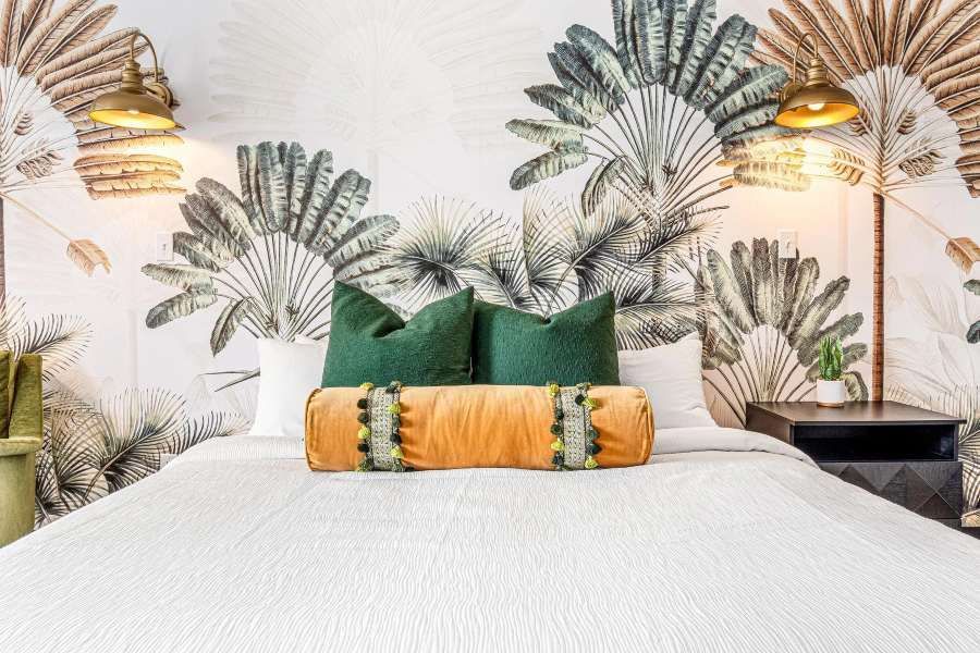 Bedroom with tropical wallpaper, green pillows, orange bolster, and brass lamps.
