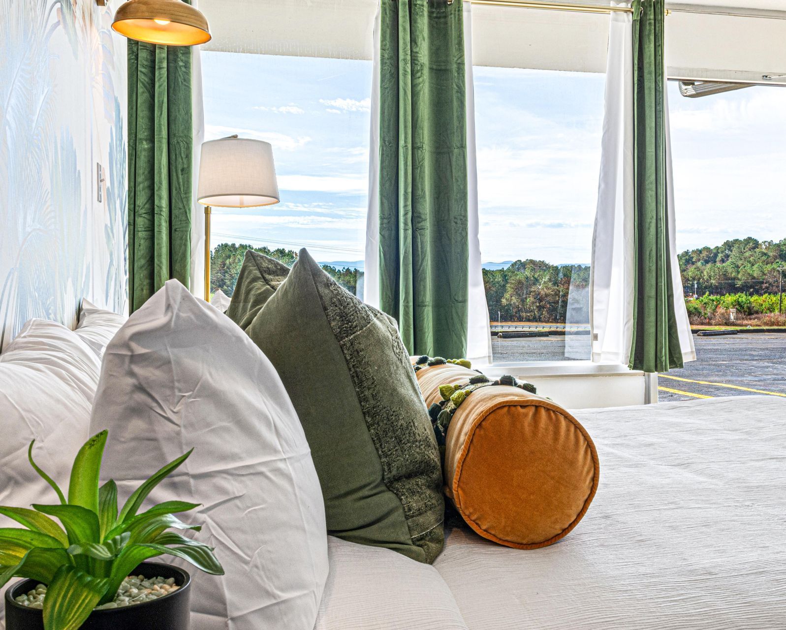 Bed with pillows, lamp, curtains, and a view of trees and sky.