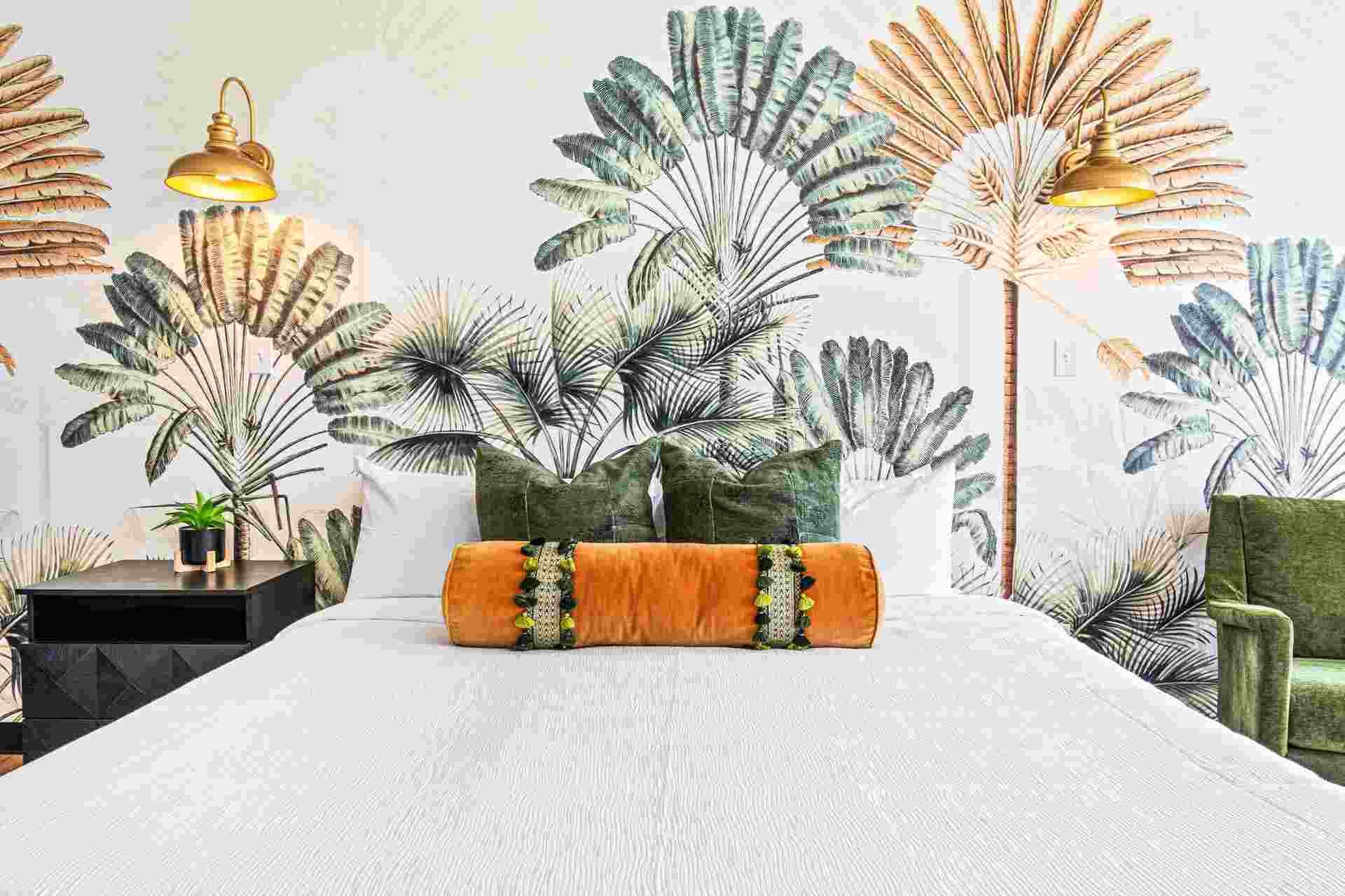 Bedroom with tropical palm mural, white bedding, orange bolster pillow, and green velvet pillows.