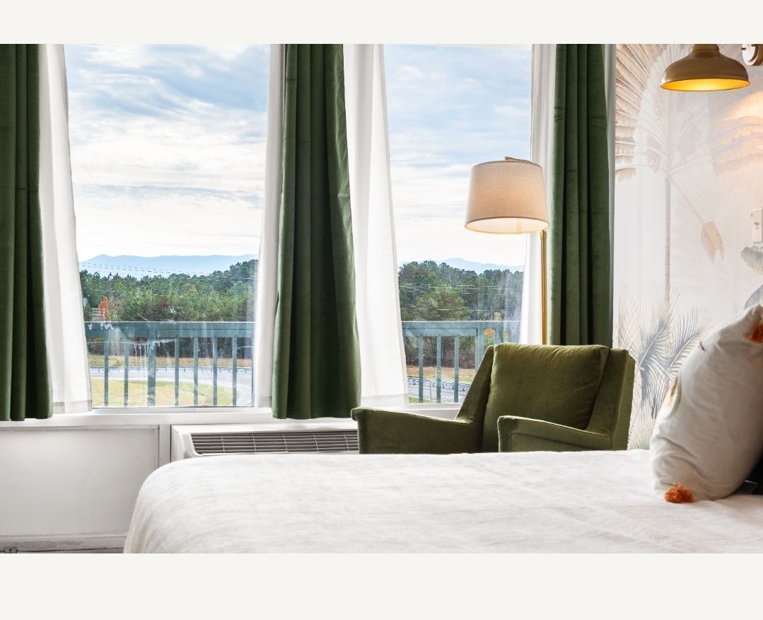 Bedroom with a bed, green chair, floor lamp, and a large window overlooking a balcony and lush scenery.