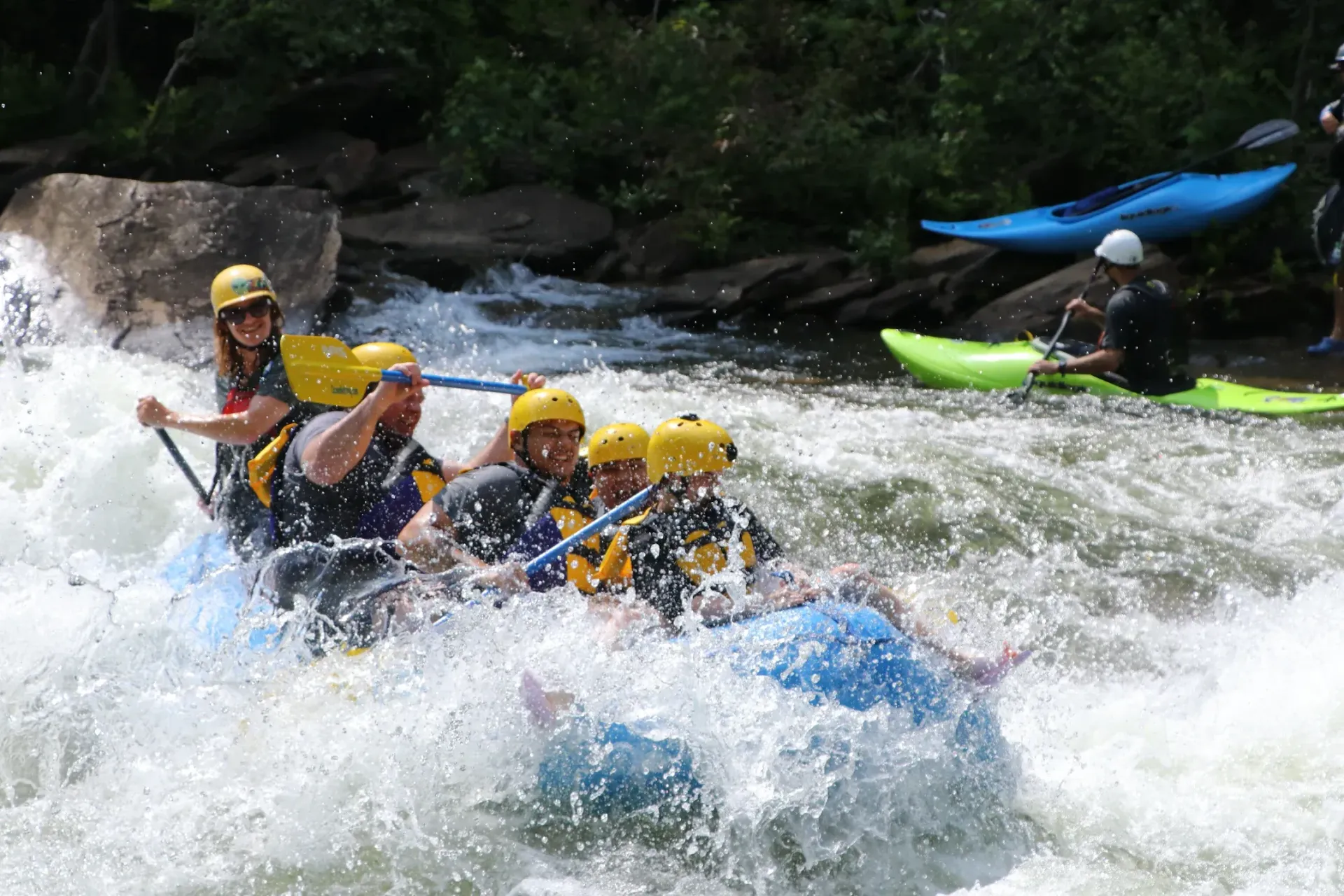 People white water rafting in a rushing river; blue raft, yellow helmets, paddles, splashing water.