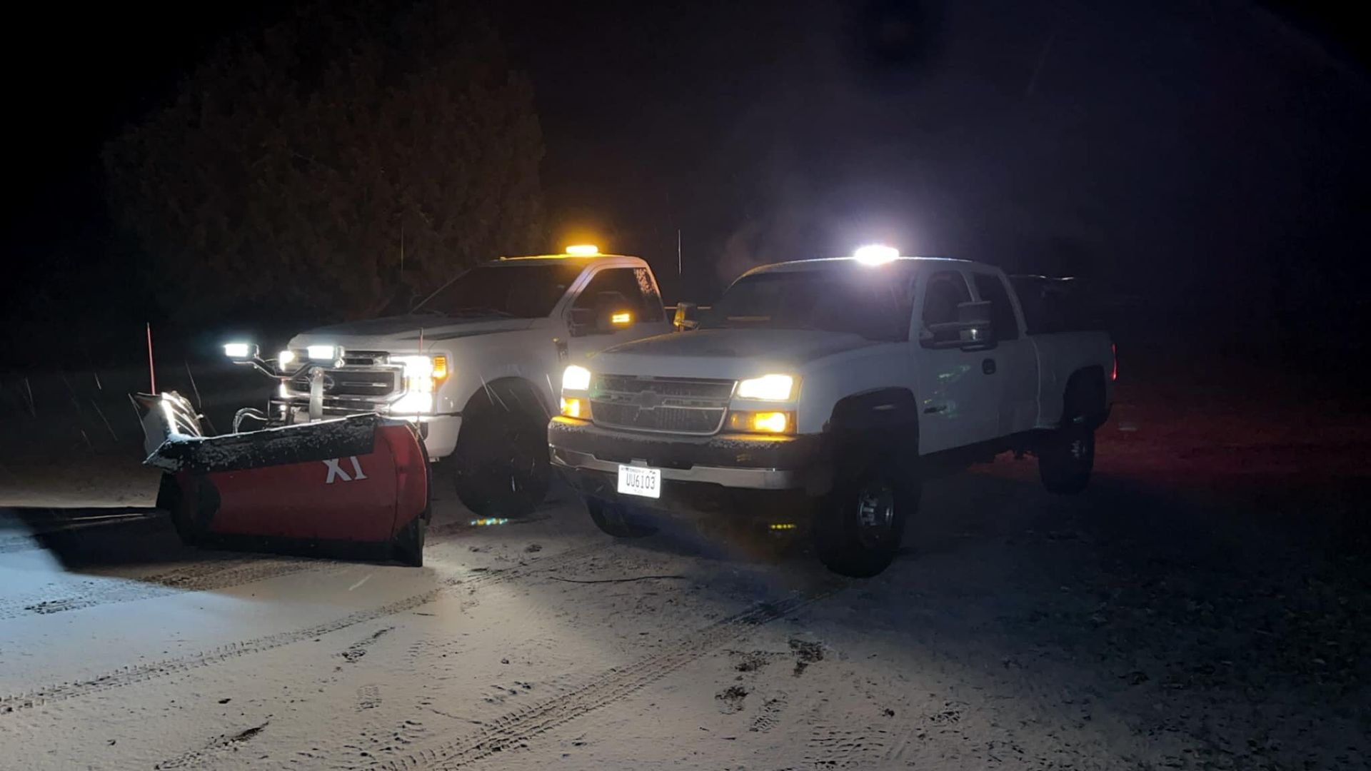 Two trucks plowing snow on a dark road.