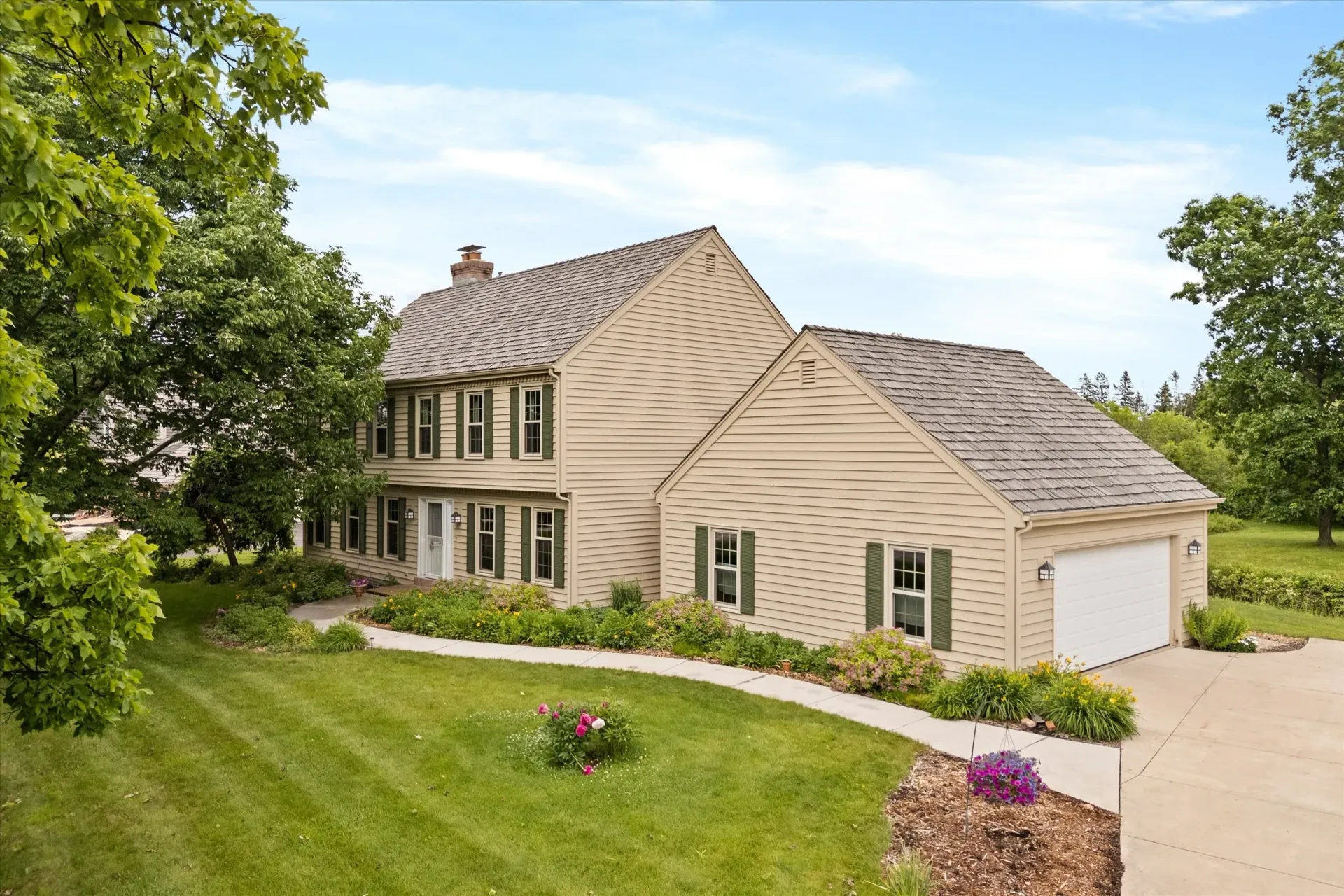 Two-story beige house with attached garage, green lawn, and trees under a blue sky