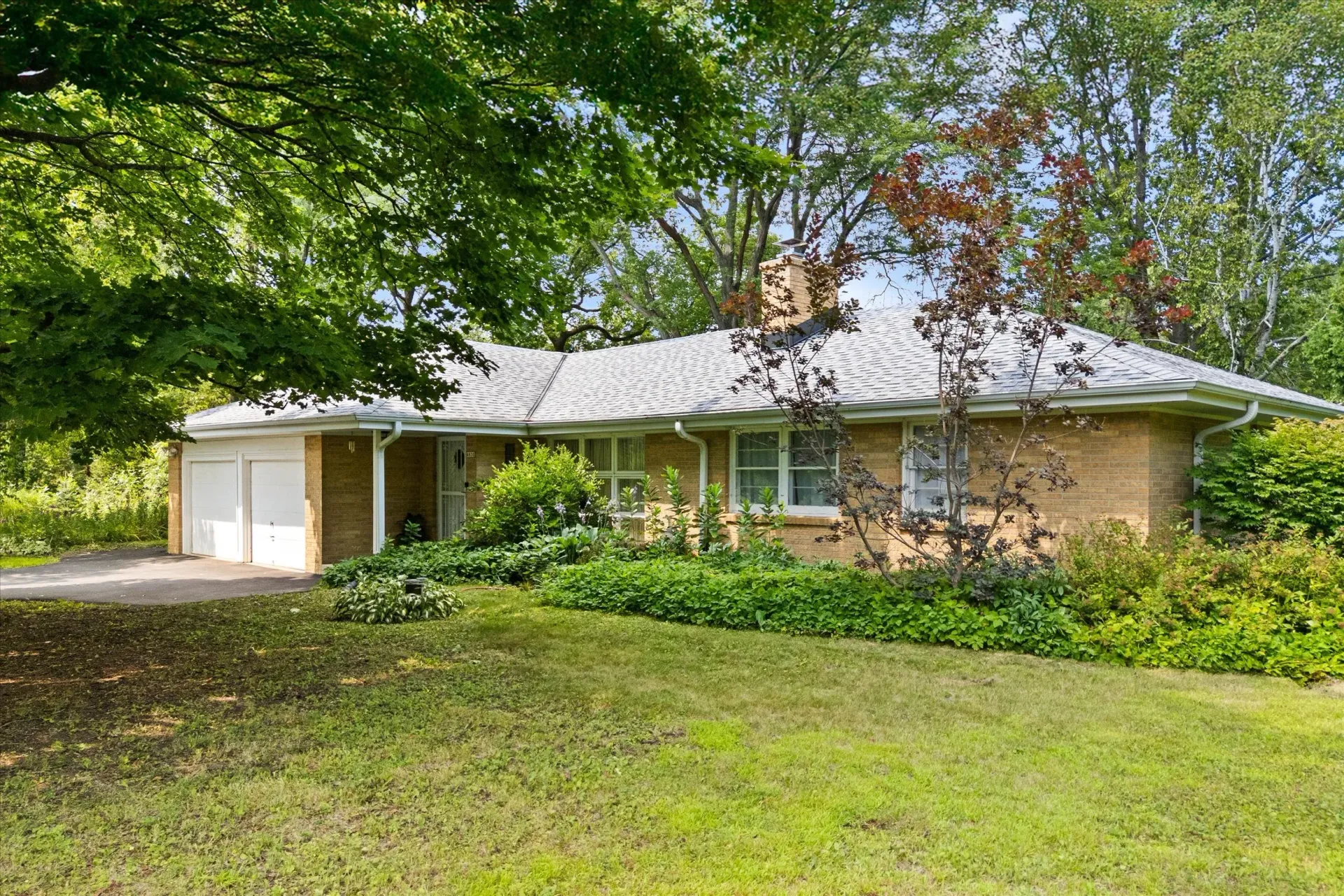 Single-story brick house with a two-car garage, surrounded by large trees and a green lawn.