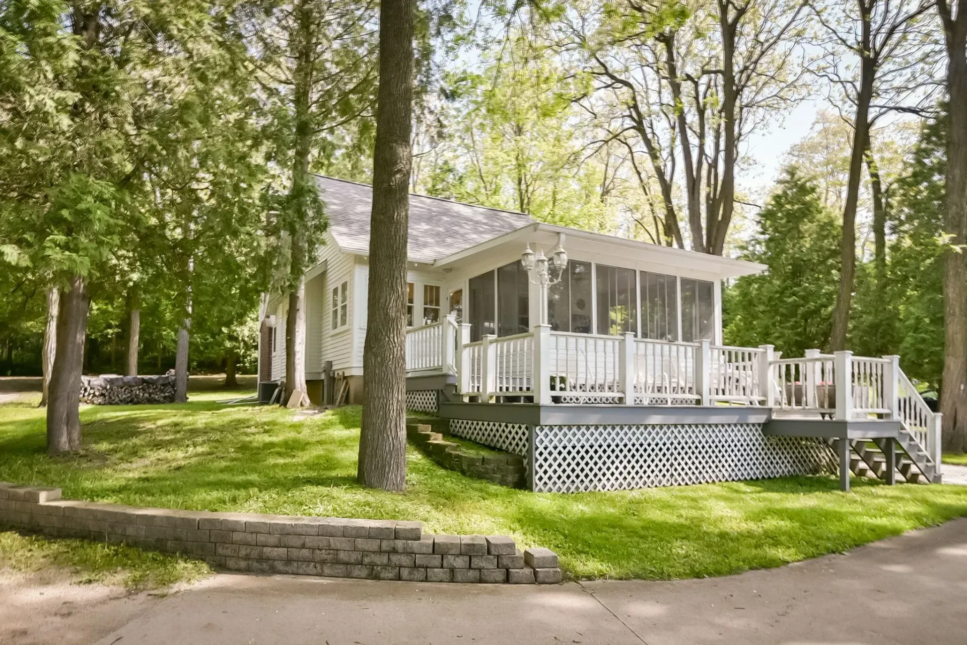 White house with wraparound porch and stone foundation among green trees and lawn.