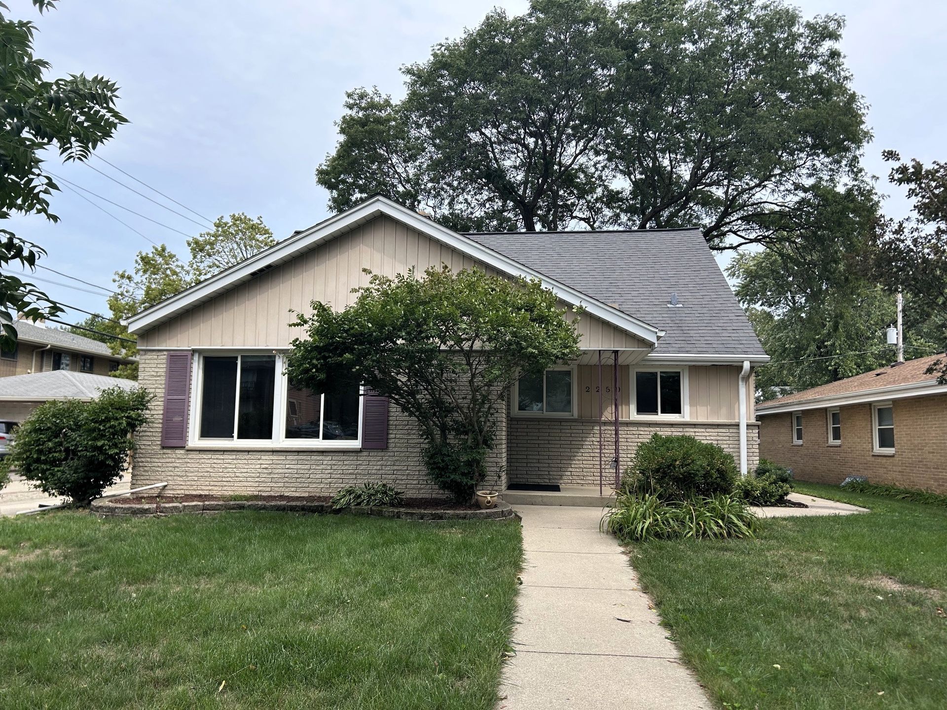 Single-story beige house with a front lawn, concrete walkway, and trees in the yard
