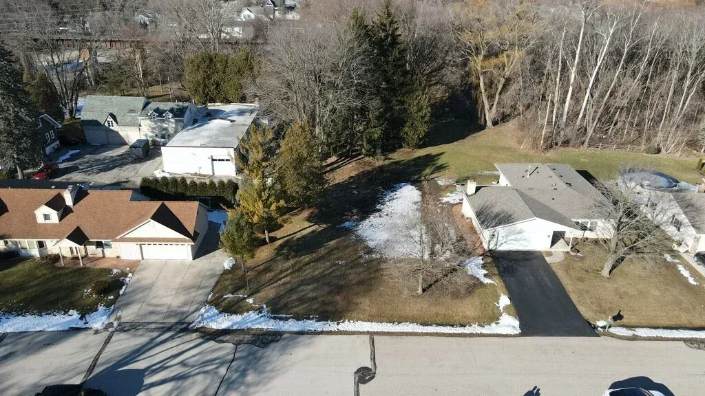 Aerial view of suburban homes, driveways, and snow-dusted yards beside a road and wooded area
