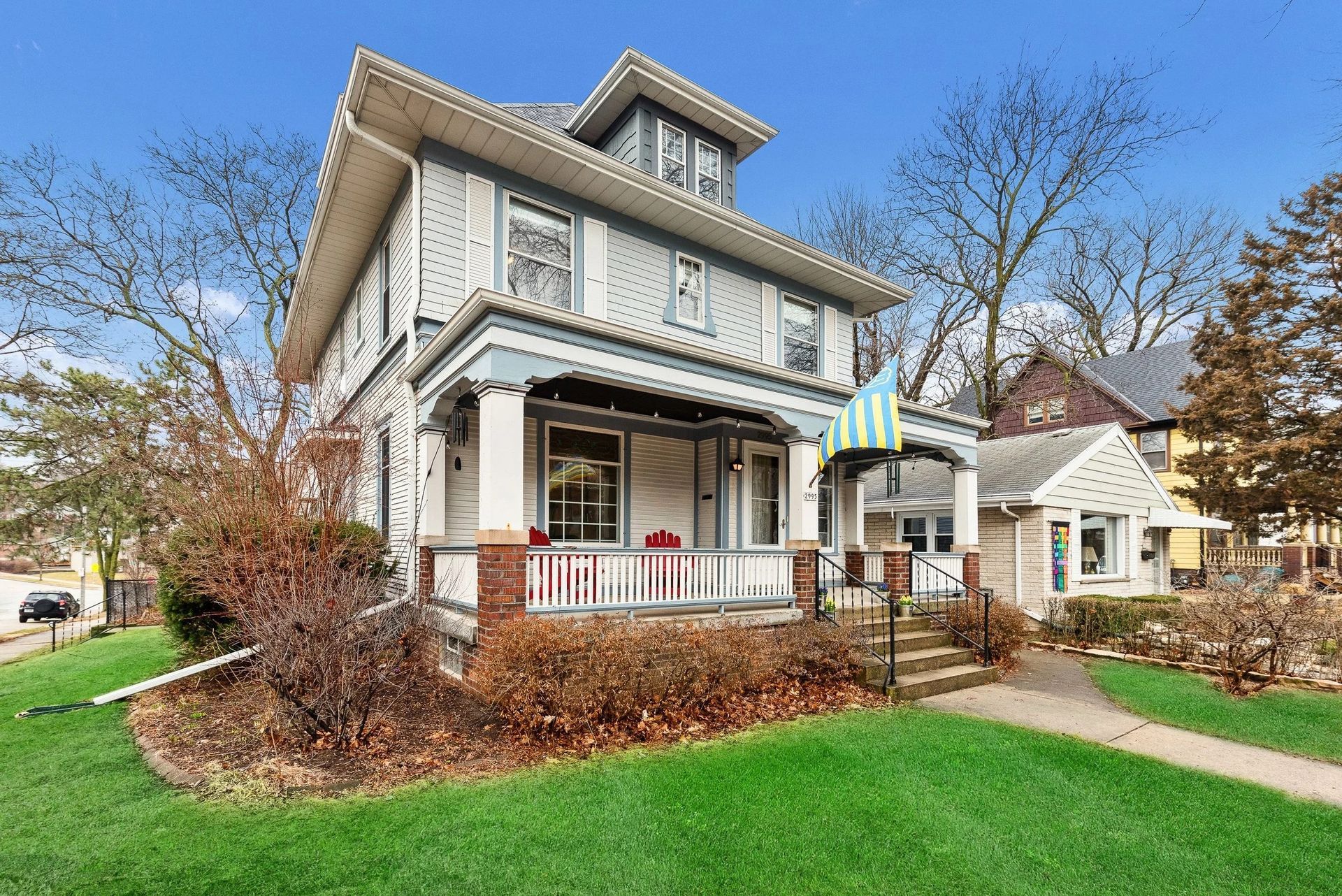 Two-story gray house with front porch, green lawn, and autumn trees under a blue sky