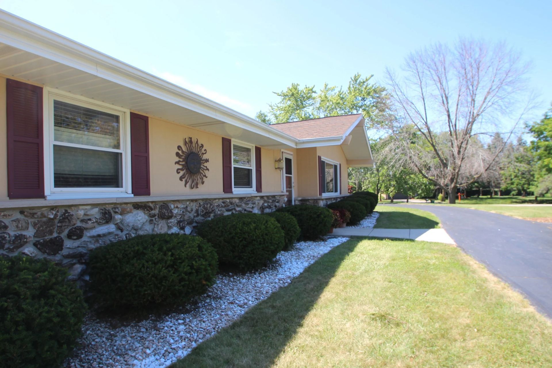 Single-story house with stone facade, manicured bushes, and a sunlit lawn beside a driveway.