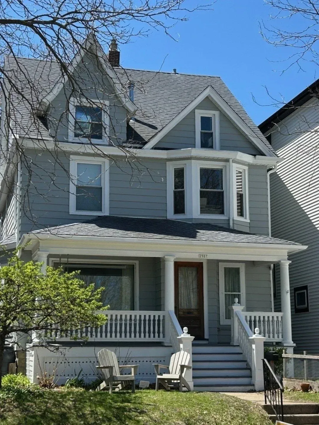 Gray two-story house with front porch, white trim, and steep gabled roof under a clear blue sky