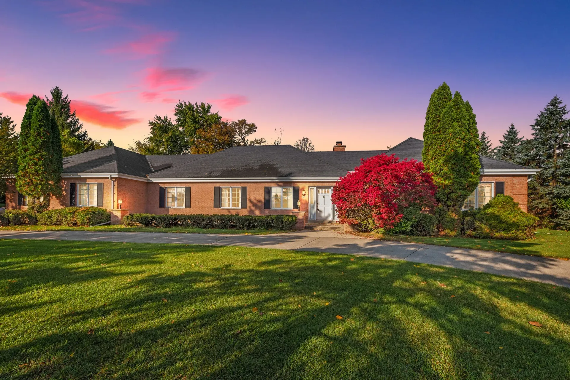 Suburban brick house at sunset with a manicured lawn and red flowering shrub under a colorful sky