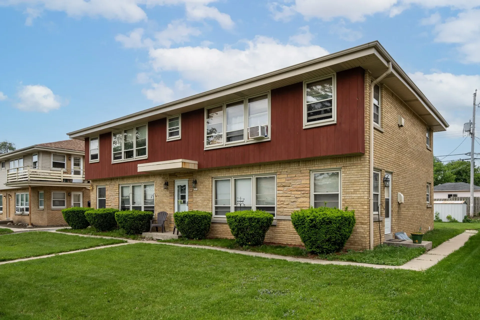 Two-story apartment building with red and tan exterior, white trim, and a green lawn under a cloudy sky