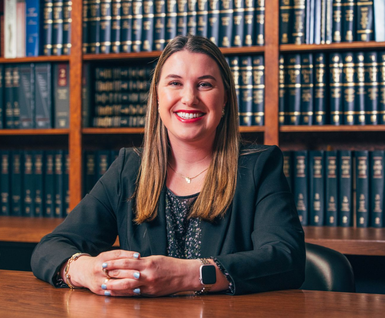 Woman in a blazer smiles at the camera, sitting at a desk with a library backdrop.