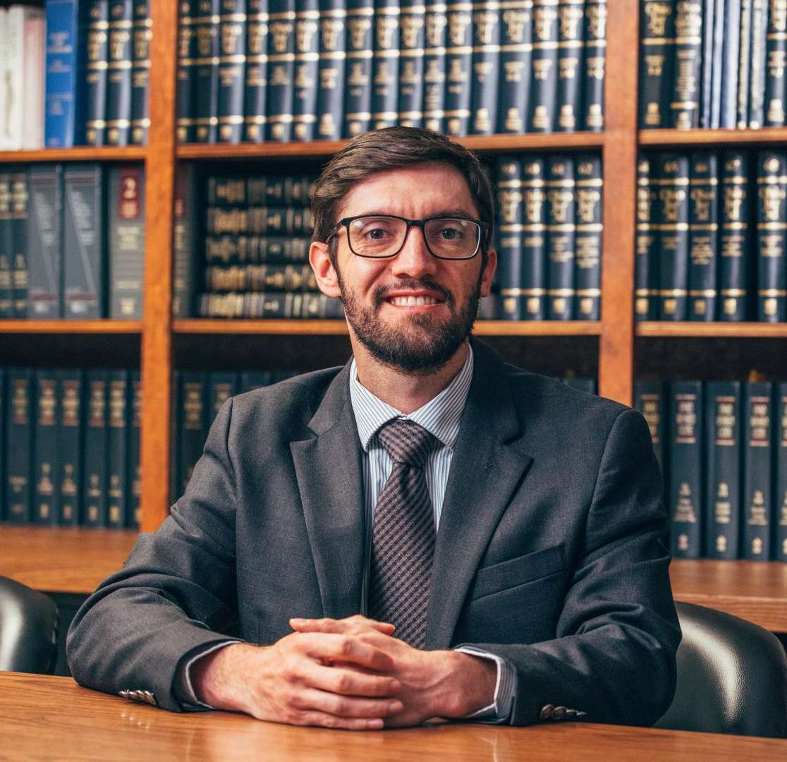 Man in suit and glasses stands in front of a bookshelf filled with books.