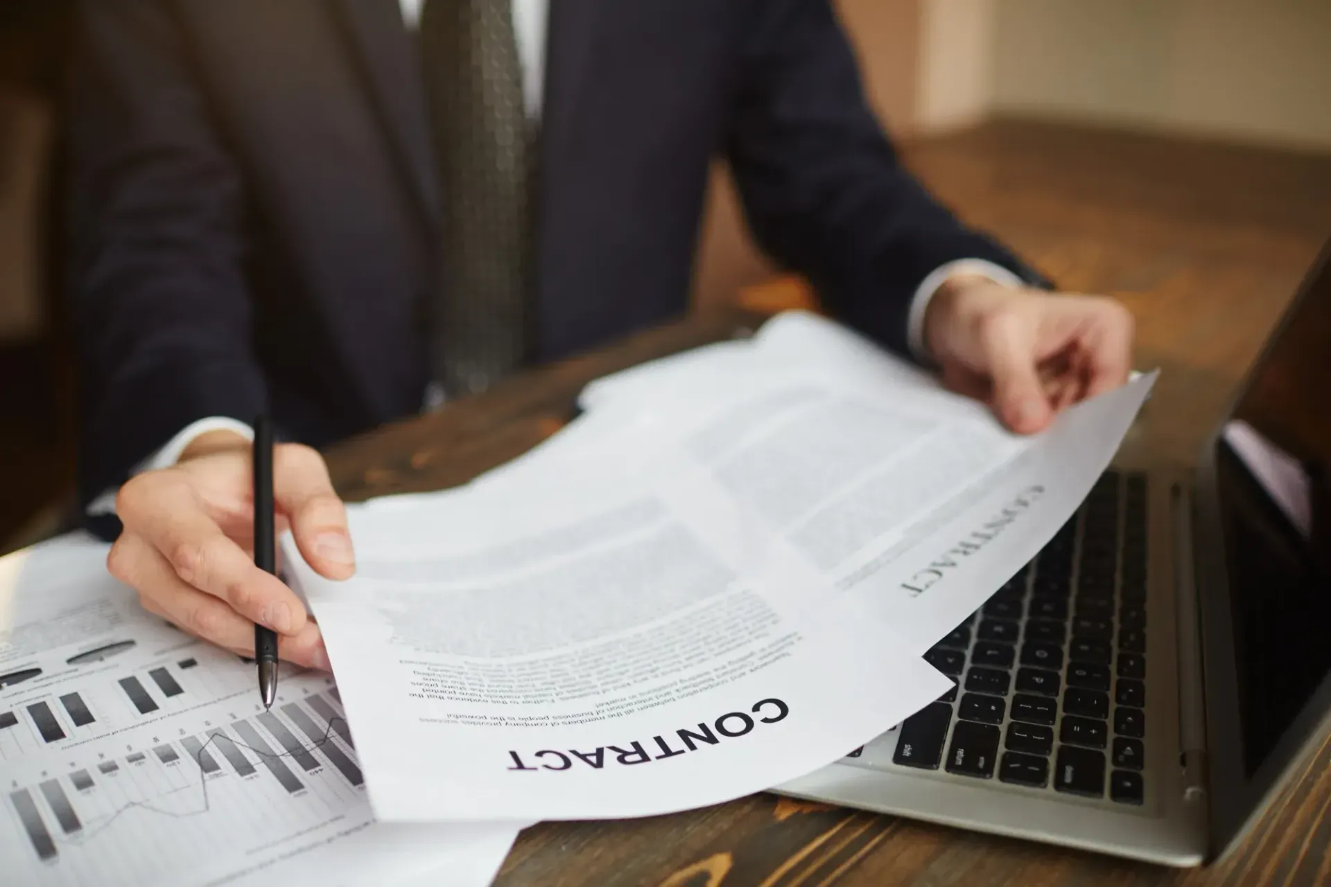 Person in suit reviewing contract documents at a desk, with a laptop and charts.