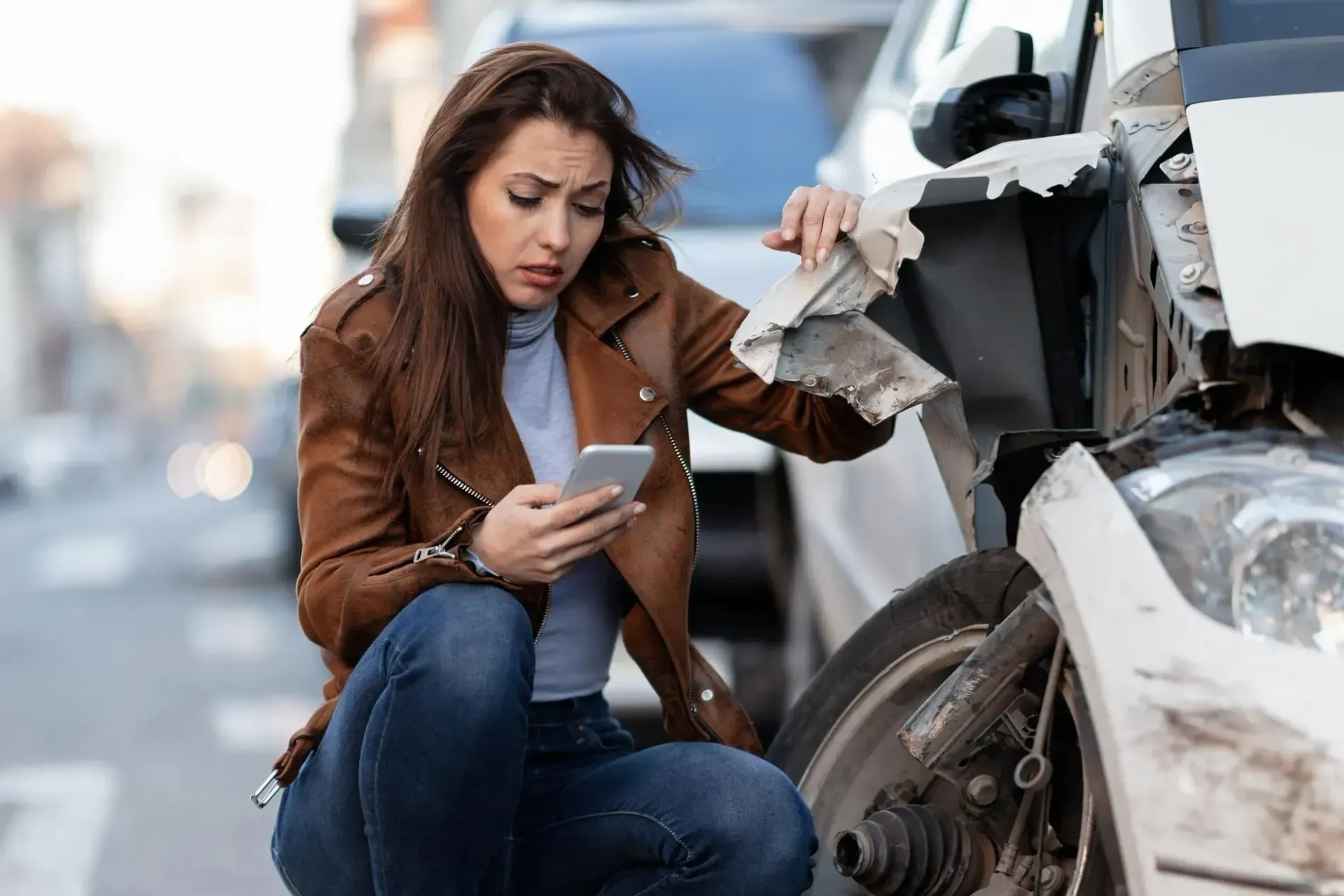 Woman looking at phone near damaged car. Brown jacket, street setting.