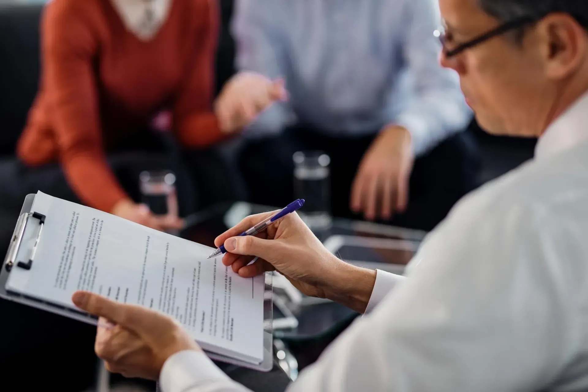 Therapist writing on clipboard; couple in background.