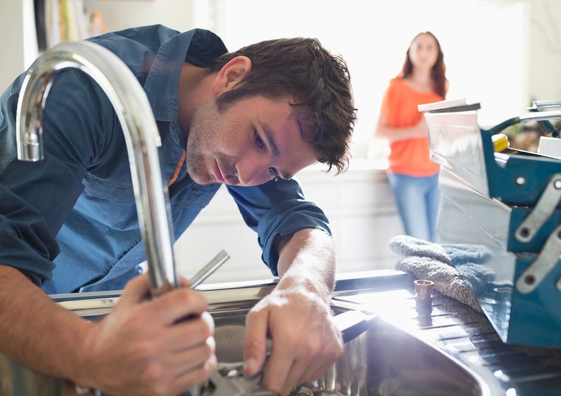 A plumber working on a kitchen sink with a blurred client looking on in the background.