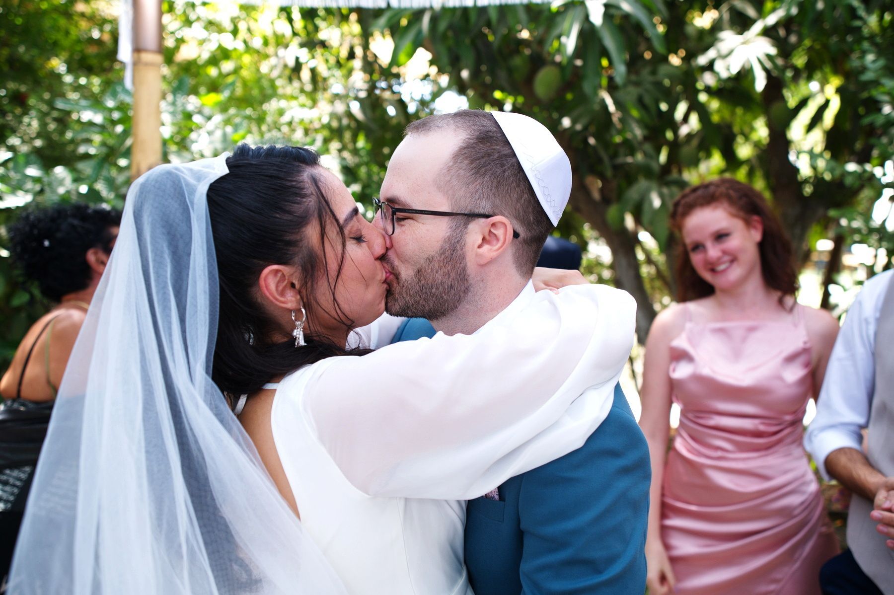 שני אנשים מתנשקים בחוץ בחתונה, כשברקע נראים אורחים וצמחייה.