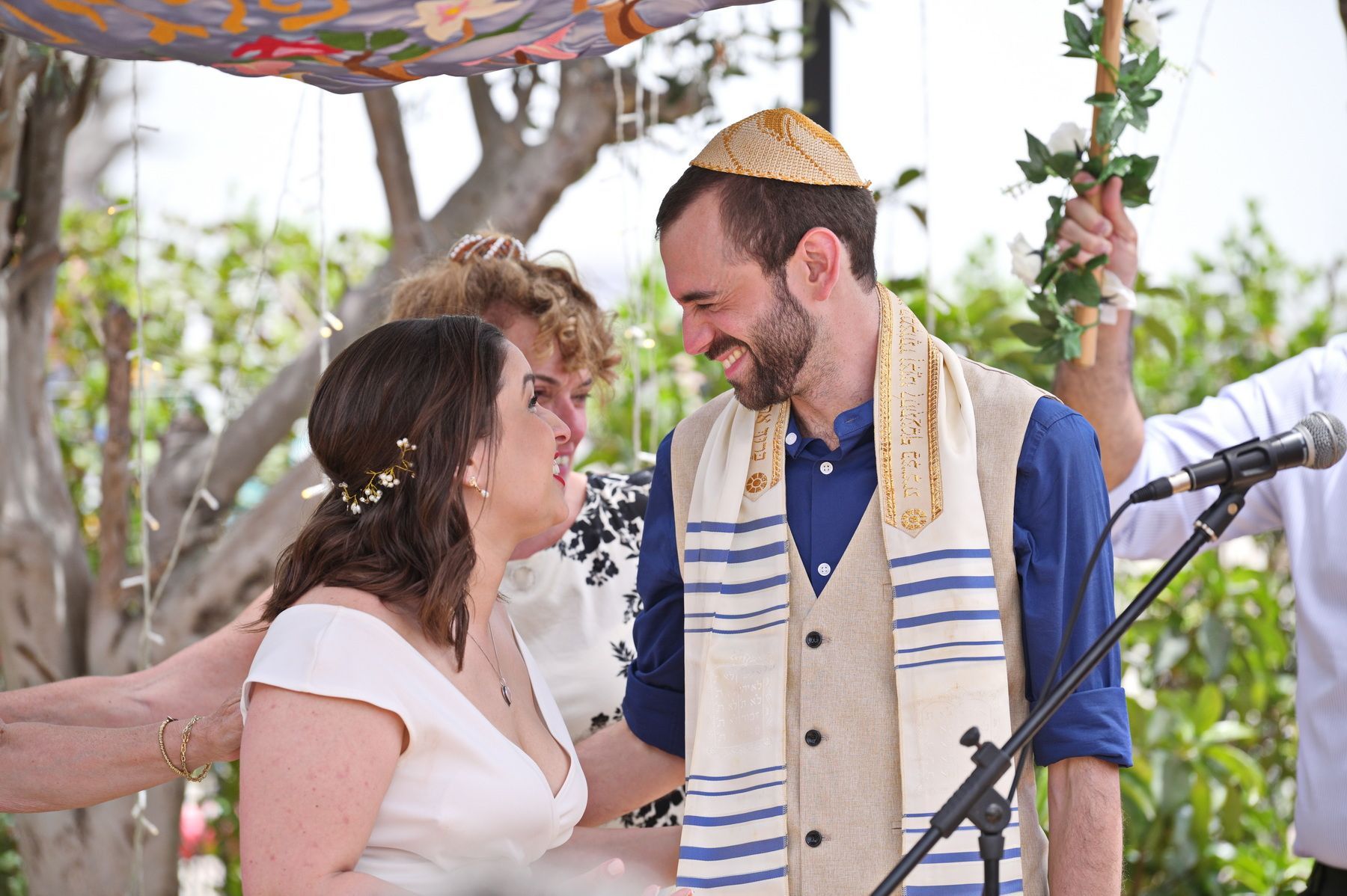 זוג מתחת לחופה במהלך טקס חתונה, מחייכים זה אל זו ליד מיקרופון.