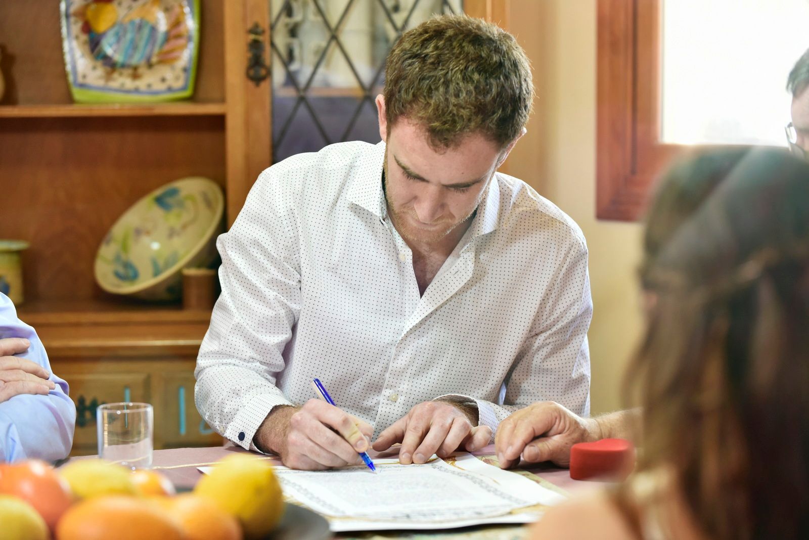 גבר כותב על ניירות ליד שולחן עץ במטבח מואר, כשפירות נמצאים בחזית התמונה - צילום חתונות - גיל דור