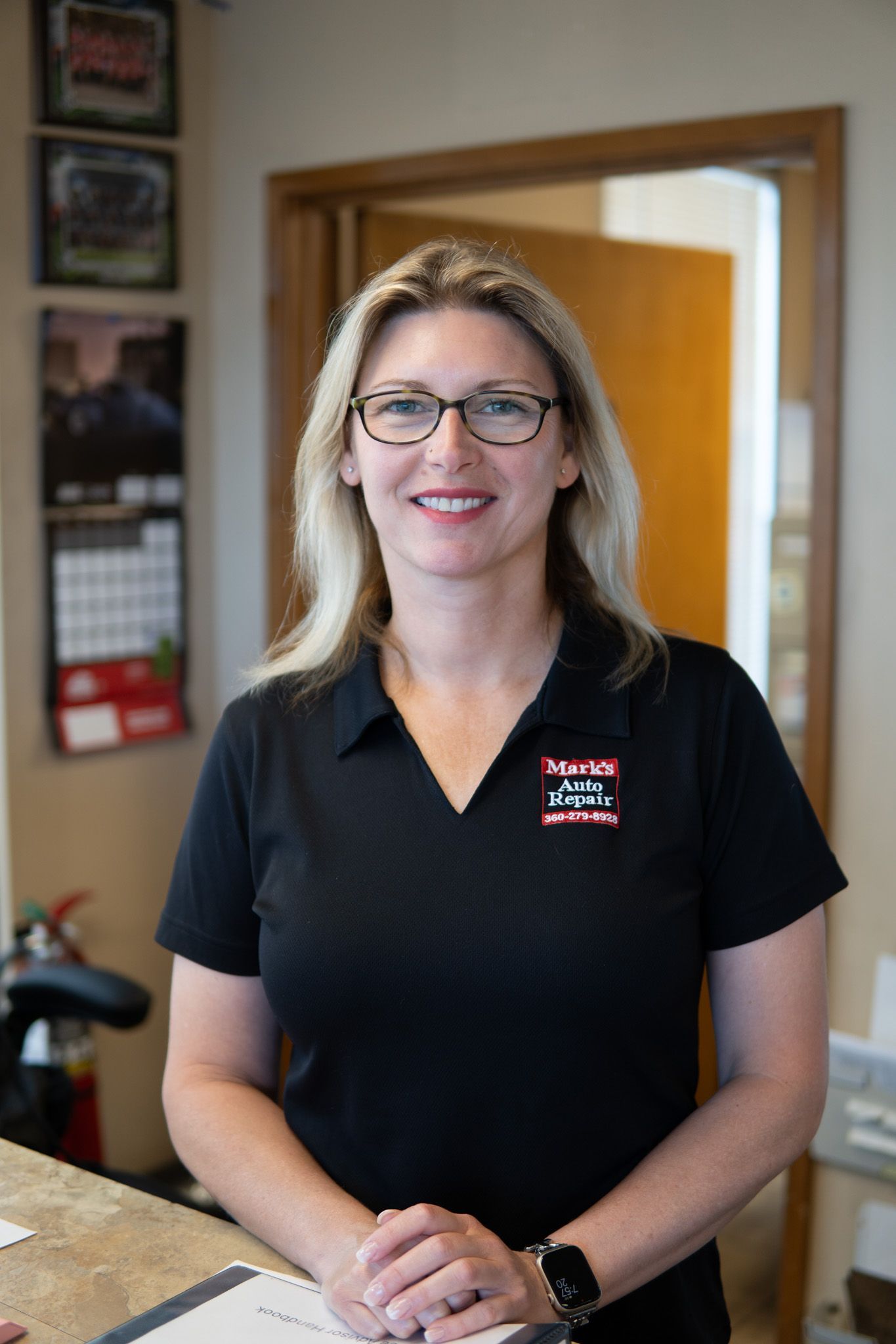 Woman with glasses and a black polo shirt smiling at a desk.