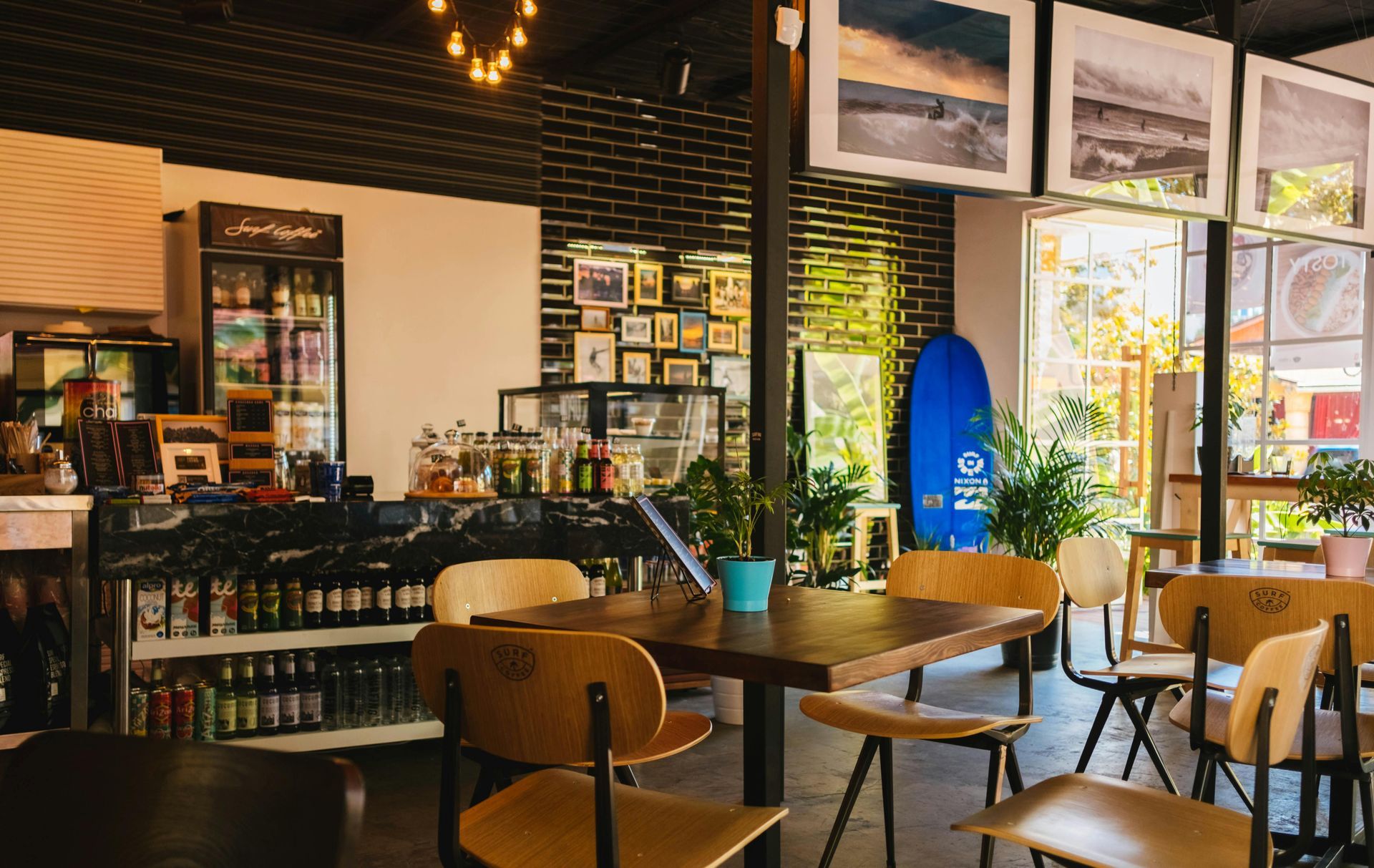 Interior of a cafe with tables, chairs, and a bar with drinks. Photos and a blue surfboard decorate the walls.