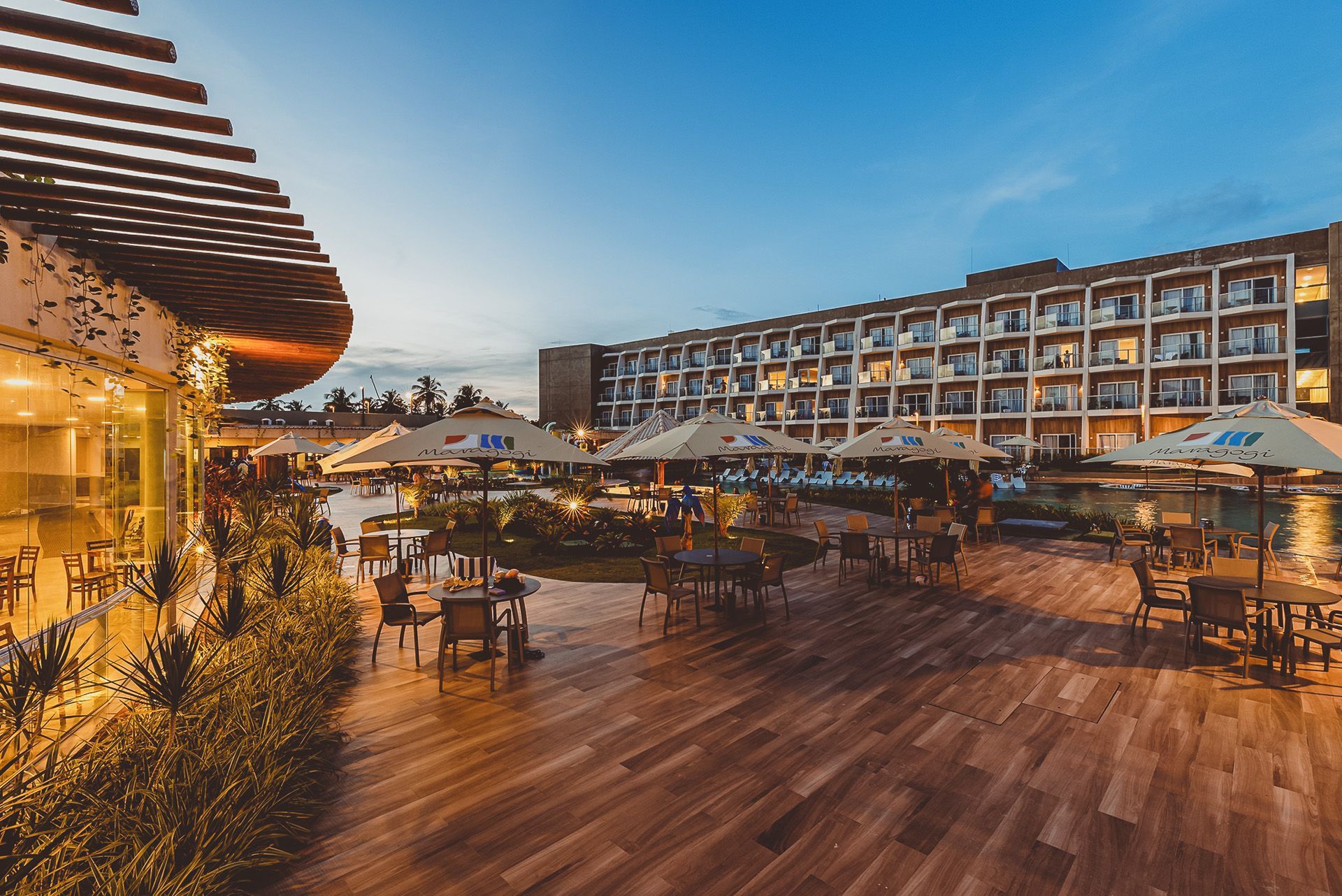 Um pátio de hotel ao ar livre com mesas, guarda-sóis e um prédio de vários andares ao anoitecer.