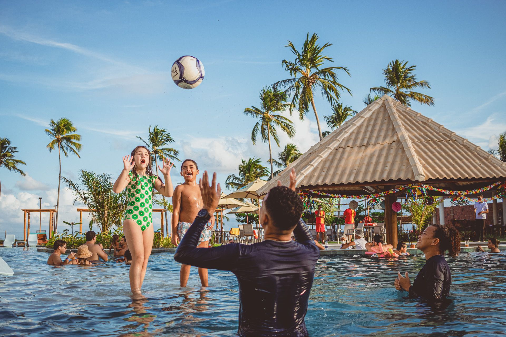 Pessoas jogando vôlei em uma piscina em um dia ensolarado com palmeiras ao fundo.