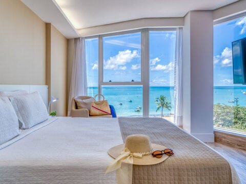 Quarto com vista para a praia; roupa de cama branca, chapéu de palha, oceano através de grandes janelas.