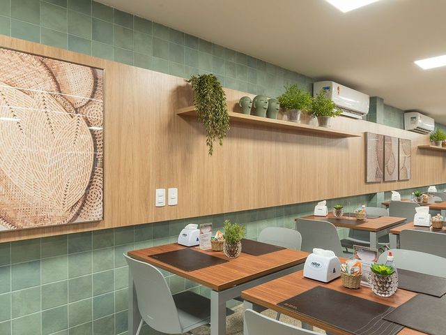 Interior do restaurante: mesas de madeira, parede de azulejos verdes, painéis de madeira com plantas e obras de arte, cadeiras cinza.